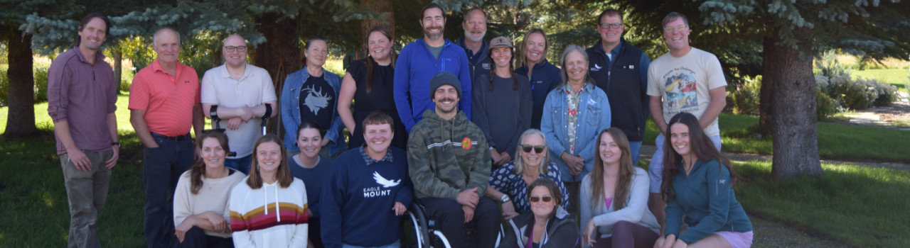 A group of 20 adults pose together outdoors on grass, some standing and others sitting, with trees in the background.